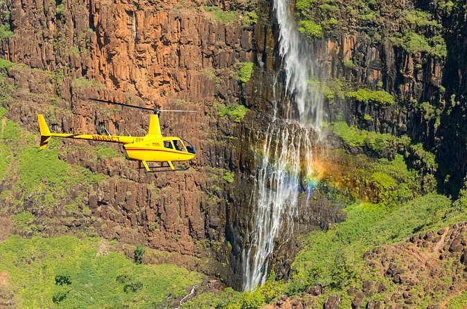 Ein kleiner Hubschrauber vor einem hohen Wasserfall an einer grünen Klippe